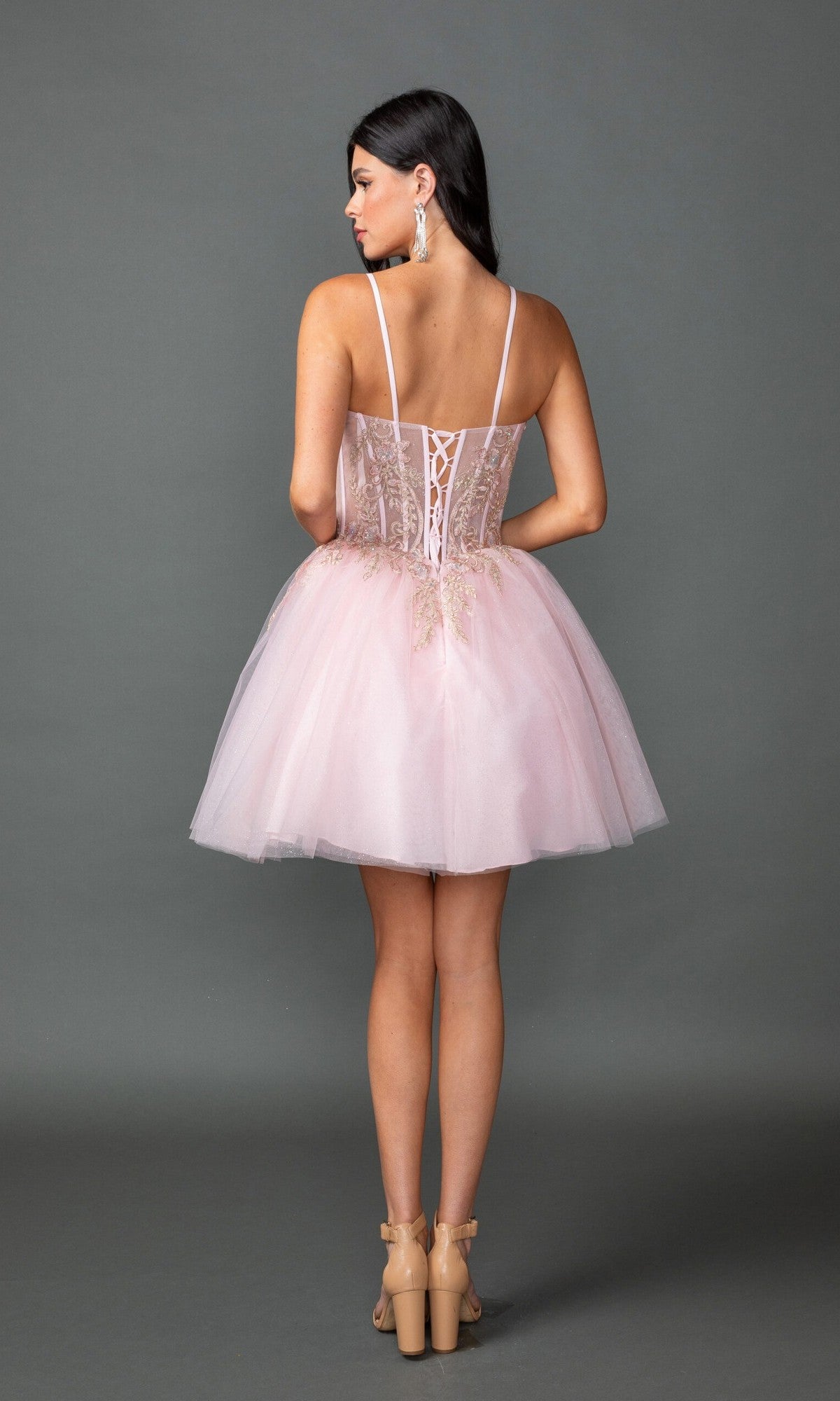 A woman faces away wearing the Dancing Queen 3424 short prom dress, featuring a light pink tulle skirt, fitted sheer corset bodice, and spaghetti straps. Her long dark hair and nude heels complete the look.